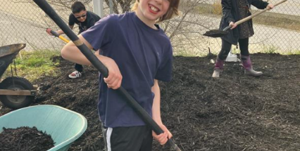 Boy in a garden holding a shovel with a wheelbarrow of soil beside him